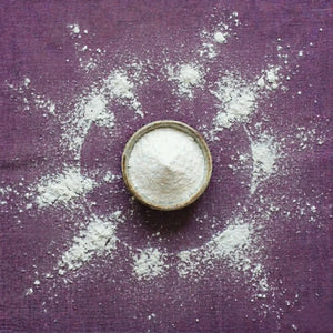 Pure micronized creatine monohydrate powder in a bowl, with white powder scattered in a sunburst pattern on a purple background, representing clean energy ingredients in Somnii.