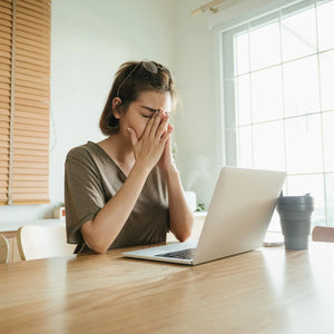 A stressed woman sitting at a desk with a laptop, covering her face in exhaustion; illustrating the maternal burnout and cognitive fatigue that Somnii wellness products are designed to support.