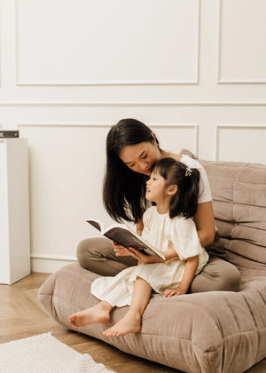 A woman with long black hair sits on a tan corduroy floor chair, holding a book and reading to a young girl in a white dress who is sitting in her lap.