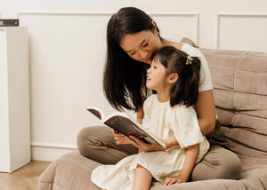 A mother reading a book to her daughter while sitting on a cozy floor sofa, representing the calm, connected presence that Somnii wellness products help mothers achieve by managing stress and fatigue.