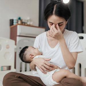 An overwhelmed mother holding her sleeping baby while pinching the bridge of her nose in exhaustion, illustrating the sensory overload and maternal fatigue addressed by Somnii wellness solutions