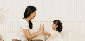 A mother and young daughter playing and high-fiving on a white sofa, representing the patient, present parenting made possible by Somnii’s maternal wellness and energy support.