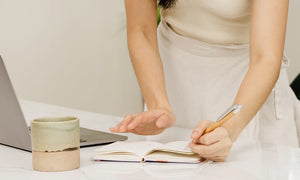 A mom writing in a journal on a marble desk next to a laptop and a ceramic mug.