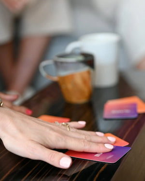 A close-up of a hand with a ring and light pink manicure selecting a Somnii Sessions card from a table, with out-of-focus coffee mugs in the background representing a cozy community gathering.