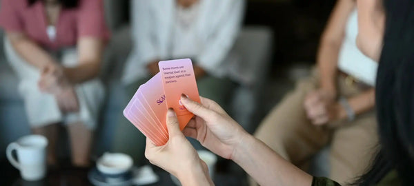 A mom holding a Somnii Sessions card while sitting with friends. The card reads: 'Some moms use mental load as a weapon against their partners.'