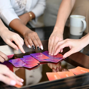 A diverse group of moms' hands reaching for Somnii Sessions cards spread out on a glossy table, demonstrating group storytelling and connection