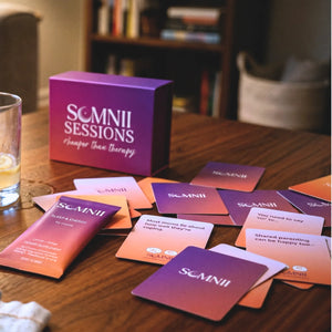 Somnii maternal wellness products on a wooden table, featuring the Somnii Sessions 'cheaper than therapy' conversation card box and Somnii's Sleep & Energy supplement sachet for moms.