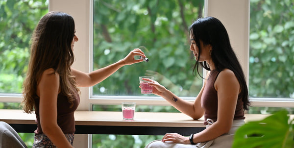Somnii founders sitting at a light wood table in front of a large window. One founder is pouring a sachet into the other's glass. 