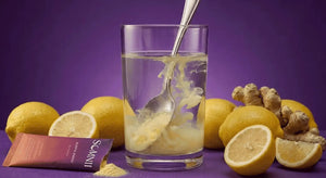 A Somnii sleep and energy supplement packet with loose powder being stirred into a glass of water by a metal spoon, surrounded by whole and halved fresh lemons and ginger root against a deep purple background.